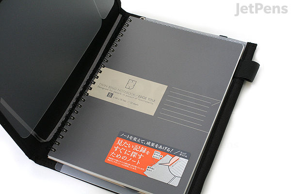 A black KOKUYO Systemic Cover ring binder notebook in A5 size with fabric cover, featuring 6 mm ruled paper inside. The notebook is open, showing the front inner cover and the first page with product information in Japanese and English.