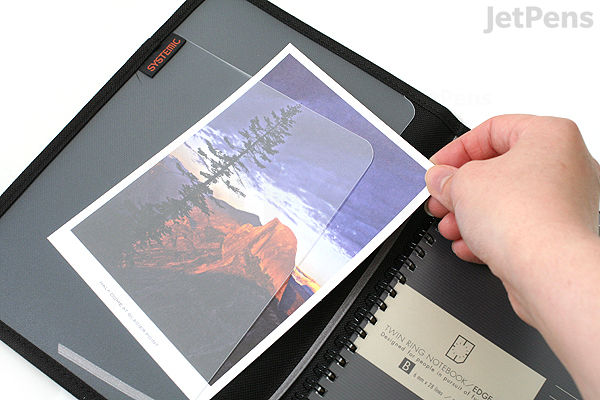 Hand holding a transparent plastic sleeve inside a black KOKUYO Systemic Cover A5 ring notebook with 6 mm ruled pages, showing a photo and a card inserted in the sleeve.