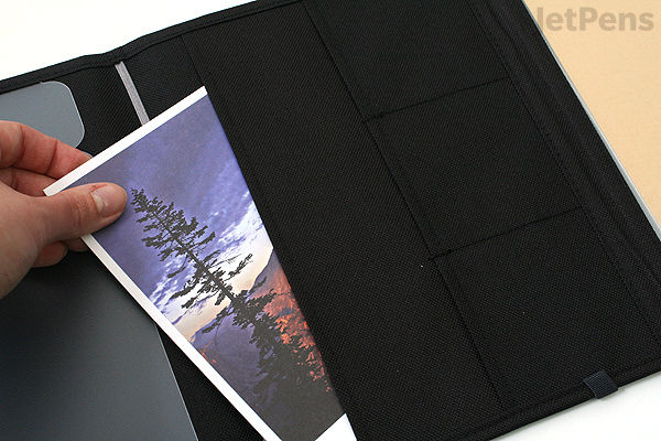 A hand placing a postcard with a scenic mountain and tree image into a black fabric cover notebook with multiple pockets, designed for A5-sized items.