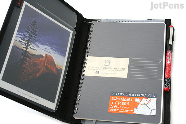 Open black KOKUYO Systemic Cover Notebook with fabric ring binder, holding an A5-sized 6mm ruled notebook and a pen, alongside a postcard featuring a mountain landscape inside the left cover pocket.