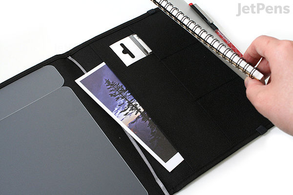 A hand flipping through the pages of a black KOKUYO Systemic Cover A5 ring notebook with fabric cover and 6 mm ruled paper, showing an inside pocket holding a bookmark and a small white item.
