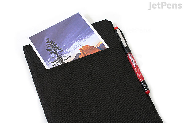 Black fabric-covered ring binder notebook in A5 size with a 6 mm ruled page partially visible, accompanied by a red and black pen clipped to the cover.