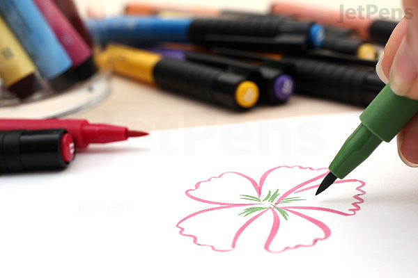 A hand using a green Faber-Castell PITT Artist Pen to draw the stem and details of a flower outlined in red on white paper, with several other colored pens scattered in the background.