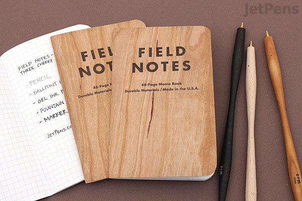 Three Field Notes Cherry Graph Memo Books with wood-grain covers, each 3.5" x 5.5" and 48 pages, shown alongside three pens on a brown surface. One notebook is open to a graph paper page with handwritten notes.