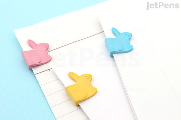 Three colorful bird-shaped paper clips in pink, yellow, and blue are attached to the edges of white and lined paper sheets on a light blue surface.