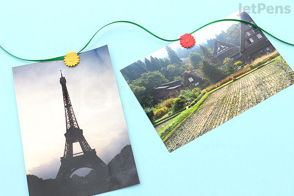Two photographs are clipped to a light blue string with colorful Midori P-51 airplane-shaped paper clips. One photo shows the Eiffel Tower against a cloudy sky, and the other depicts a scenic landscape with terraced fields and mountains.