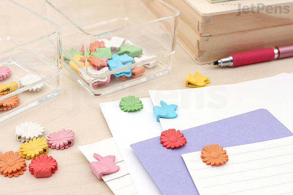 Colorful paper clips shaped like airplanes and gears scattered on a white desk next to a purple envelope, a pen, and clear storage containers.