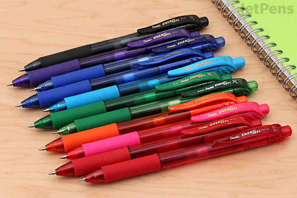 A row of nine Pentel EnerGel-X gel pens with 0.7 mm conical tips, arranged diagonally on a wooden surface. The pens are in various colors including black, purple, blue, green, and red. A green spiral notebook is partially visible in the background.