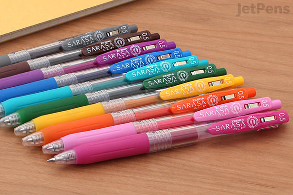A set of 14 Zebra Sarasa Clip gel pens with 0.5 mm tips arranged diagonally on a wooden surface, showcasing a variety of vibrant colors including purple, blue, green, yellow, orange, and pink.