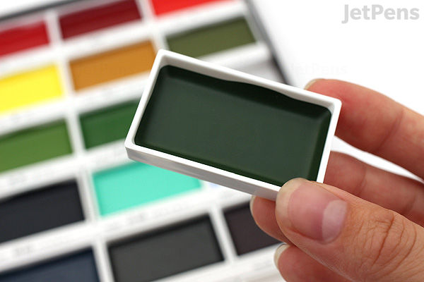 A hand holding a single rectangular pan of dark green watercolor paint from the Kuretake Gansai Tambi 24-color palette, with the full palette visible blurred in the background.