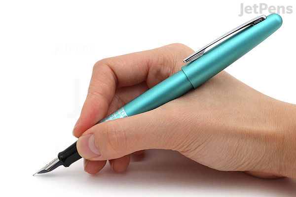 A hand holding a turquoise Pilot Metropolitan Retro Pop fountain pen with fine nib, poised for writing. The pen has a sleek, metallic finish with small dot patterns.