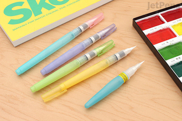 Set of five Kuretake Water Brush pens with colored caps arranged on a wooden surface next to a watercolor palette and a sketchbook.