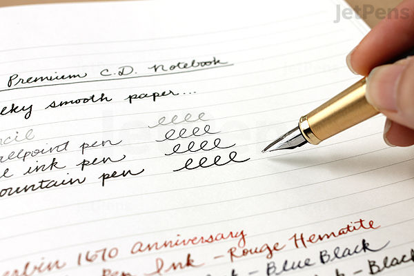 A hand holding a gold and black fountain pen writing neat cursive text on a white Apica Premium C.D. Notebook page with 5 mm graph lines. The text includes pen names and ink colors written in black and red ink.