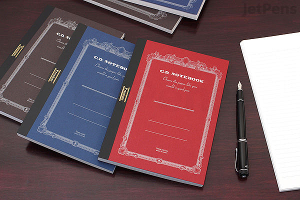 A5 Apica Premium C.D. Notebooks with 5 mm graph paper in blue, red, and brown covers, arranged on a wooden desk next to a pen and an open blank notebook.