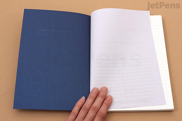 An open Apica Premium C.D. Notebook A5 size with 7 mm ruled pages, showing a blue cover on the left and lined white paper on the right, held by a hand on a light wooden surface.