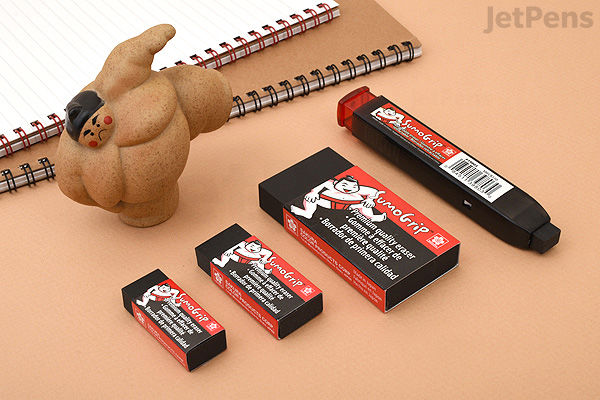 A set of Sakura Sumo Grip retractable erasers in four different sizes, each with black and red packaging featuring a sumo wrestler illustration. The erasers are displayed on a beige surface next to a sumo wrestler figurine and a spiral notebook.