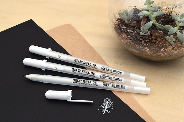 Three white Sakura Gelly Roll Classic gel pens with 0.8 mm tips are arranged on a wooden surface next to a black notebook and a small glass bowl containing succulents. One pen has its cap removed and placed nearby.