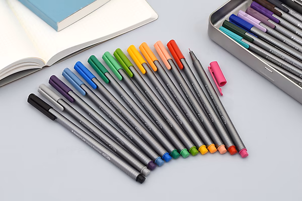 Set of ten Staedtler Triplus Fineliner pens with 0.3 mm tips, arranged in a row on a light surface, next to an open notebook and a box containing additional colored pens.