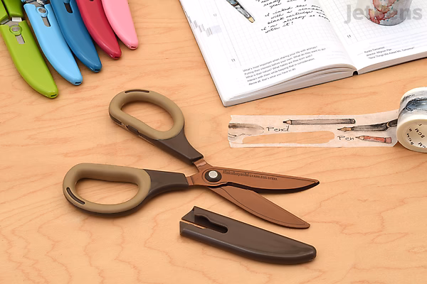 Nakabayashi Sakutto Cut Hikigiri scissors with brown handles and a protective black sheath, placed on a wooden surface near an open book and stationery items.