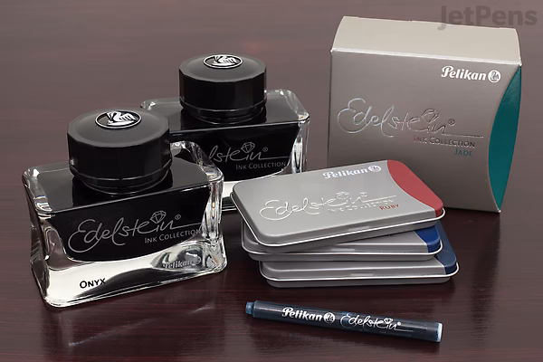 Two glass bottles of Pelikan Edelstein Ruby ink, one open with a visible red ink reservoir, placed on a dark surface next to a stack of color swatch cards and a small ink sample vial, with the product box in the background.