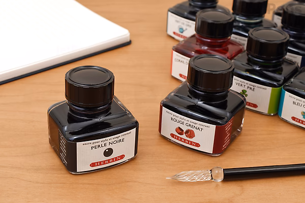 A 30 ml square glass bottle of Herbin Ambre de Birmanie (Amber Gold of Burma) ink with a black cap, placed on a wooden surface next to a glass dip pen and several other ink bottles in the background.
