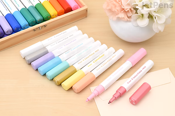 Set of six Pilot Juice Paint Markers with extra fine metallic tips, displayed uncapped in pastel colors on a wooden surface next to a white flower vase and an open notebook.