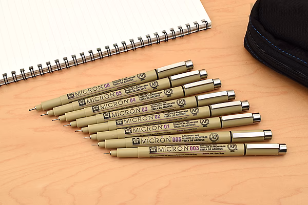 A set of seven Sakura Pigma Micron pens, size 003 (0.15 mm), with black ink, arranged in a row on a wooden surface. A spiral-bound sketchbook and a black zippered case are partially visible in the background.