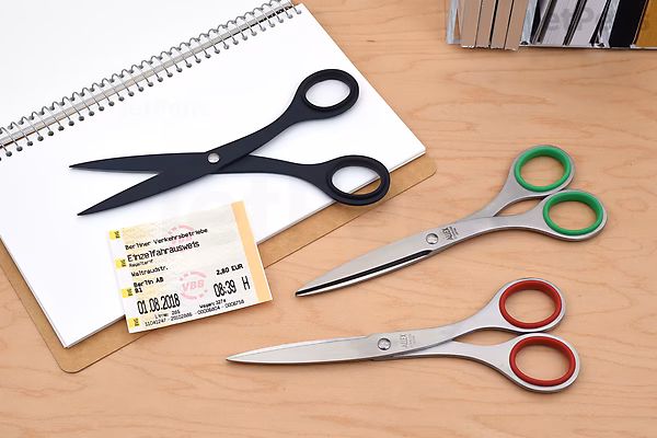 Three pairs of scissors on a wooden desk: one black pair labeled Allex S-165 Office Scissors resting on an open notebook, and two silver pairs with green and red handles lying nearby next to a business card.