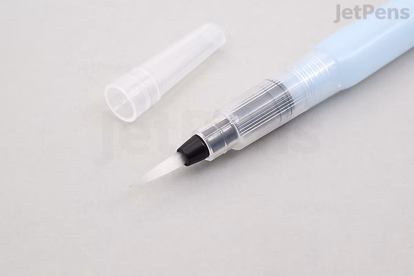 Close-up image of a Sakura Koi Watercolor Field Sketch Box Set water brush with a transparent cap placed beside it on a white surface. The brush tip is black and designed for watercolor painting.