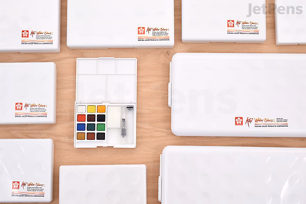 Top-down view of multiple Sakura Koi Watercolor Field Sketch Box Sets arranged on a wooden surface, featuring a compact 36-color palette and a water brush inside one open box.