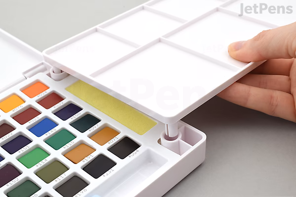 A hand lifting the white mixing palette lid of a Sakura Koi Watercolor Field Sketch Box Set, revealing a 48-color watercolor palette and a water brush inside.