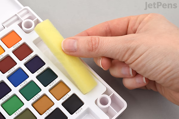 A hand holding a yellow pastel crayon above a watercolor palette with 48 vibrant color pans arranged in rows. The palette is labeled "Sakura Koi Watercolor Field Sketch Box Set."
