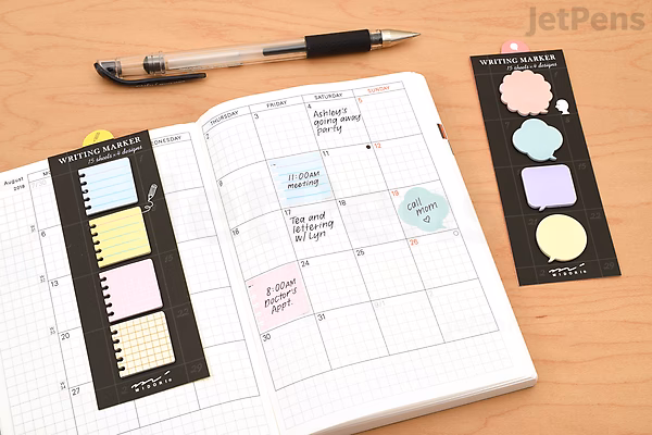 Open planner with a monthly calendar view, featuring pastel-colored Midori Writing Marker Monthly Block Sticky Notes in various shapes and sizes used for reminders. Two black sticky note sheets and a black pen lie nearby on a wooden surface.