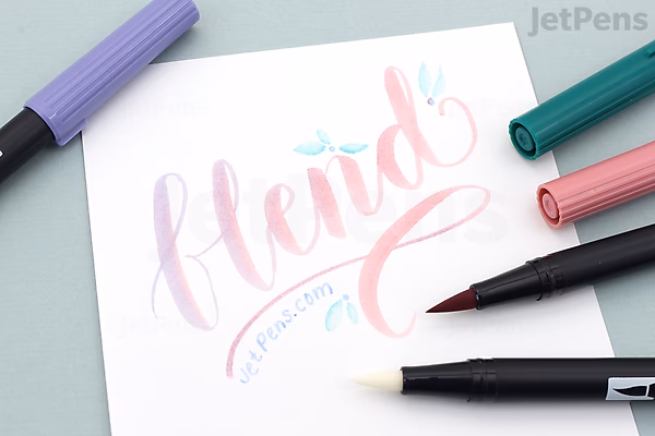 A hand-lettered word "blend" written in pink and purple shades on white paper, surrounded by four Tombow Dual Brush Pens in pastel colors, with one pen's brush tip touching the paper.
