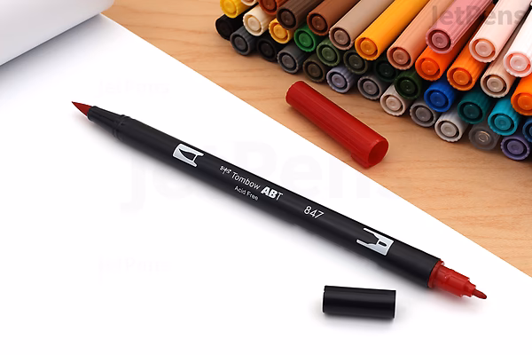 A Tombow Dual Brush Pen with a red tip lies uncapped on a white surface, next to its black cap. In the background, a set of 20 colorful dual brush pens is arranged in a wooden case.
