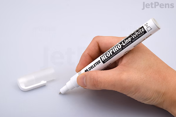 A hand holding a Deleter Neopiko Line White Drawing Pen with a 0.5 mm tip, positioned above a white surface. The pen's cap is off and placed nearby.