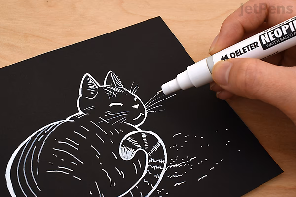 A hand using a Deleter Neopiko Line White Drawing Pen (0.5 mm) to draw white lines and details on a black paper illustration of a cat.