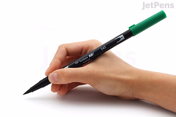 A hand holding a Tombow Dual Brush Pen with a green cap, positioned as if ready to write or draw.