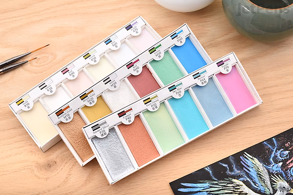 Six-color set of Boku-Undo Gansai Watercolor Palette in metallic shades, arranged in a white plastic tray on a wooden surface, with a paintbrush and a partially visible watercolor painting nearby.