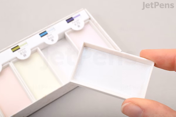 A hand holding the lid of a Boku-Undo Gansai Watercolor Palette - Aurora, showing six rectangular watercolor pans in a white plastic case with color labels above each pan.