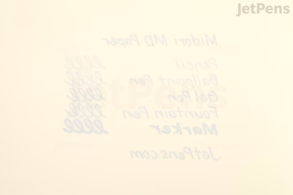 Close-up of an open Midori MD Notebook Journal in A5 size, showing a cream-colored page with handwritten ink test samples in various colors and pen types, demonstrating ink flow and shading.