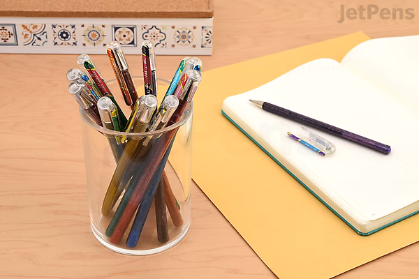 Clear container holding various colored pens and pencils on a wooden desk next to an open notebook and a yellow folder, with a decorative border on the wall in the background.