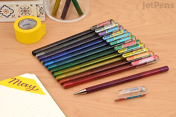 Set of eight Pentel Hybrid Dual Metallic Gel Pens, 1.0 mm tip, arranged on a wooden surface with one pen uncapped. Nearby are a notebook, yellow tape, and colored pencils in a cup.