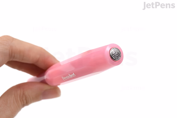 A hand holding a pink Kaweco Frosted Sport fountain pen with a broad nib, shown capped and viewed from the top end. The pen has a frosted, translucent finish.