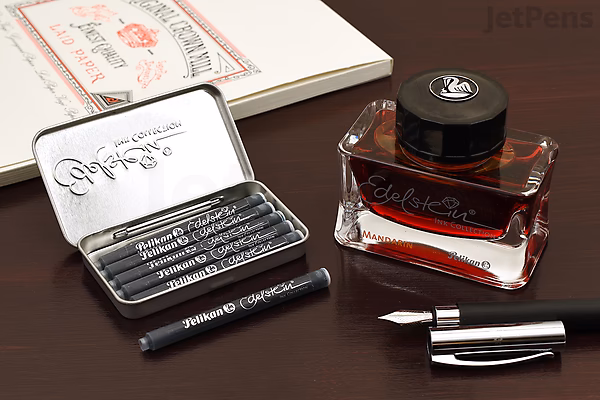 A 50 ml bottle of Pelikan Edelstein Ruby red ink with a black cap, placed on a dark wooden surface next to a disassembled black fountain pen and a small metal case containing three black ink cartridges. A partially visible notebook is in the background.