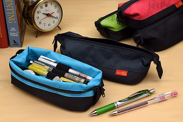 Nomadic PW-11 black boat-shaped pen case open on a desk, showing pens inside. Another pen case and two pens lie nearby, with a clock and books in the background.
