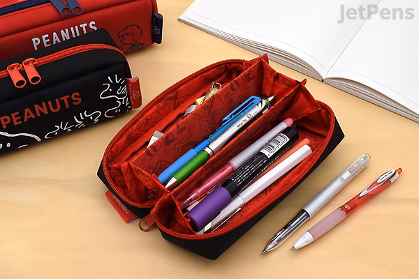 Open black Kamio Japan Paco-Tray Peanuts pen case with red interior, containing various pens and markers, placed on a wooden desk next to a notebook and other Peanuts-themed pen cases.