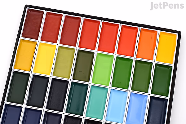 A close-up photo of a Kuretake Gansai Tambi watercolor palette containing 48 vibrant paint colors arranged in rows within a white plastic case. The colors include various shades of red, orange, yellow, green, blue, and black.