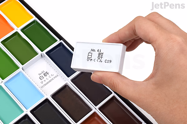 A hand holding a small white watercolor pan labeled "No. 41" above an open Kuretake Gansai Tambi watercolor palette containing 48 vibrant colors arranged in rows.