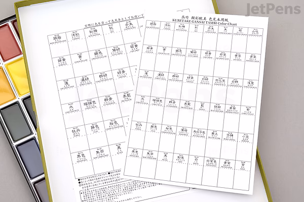 Three printed color mixing charts with numbered color swatches are placed on top of an open Kuretake Gansai Tambi 48-color watercolor palette, showing a variety of vibrant paint pans arranged in rows.
